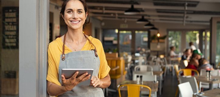 Portrait of happy woman standing at doorway of her store holding digital tablet. Cheerful mature waitress waiting for clients at coffee shop. Successful small business owner in casual clothing and grey apron standing at entrance and looking at camera.