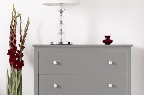 Three drawer gray cabinet with a lamp on and a vase of burgundy gladiolas by in a glamour furniture showroom interior. Real photo.