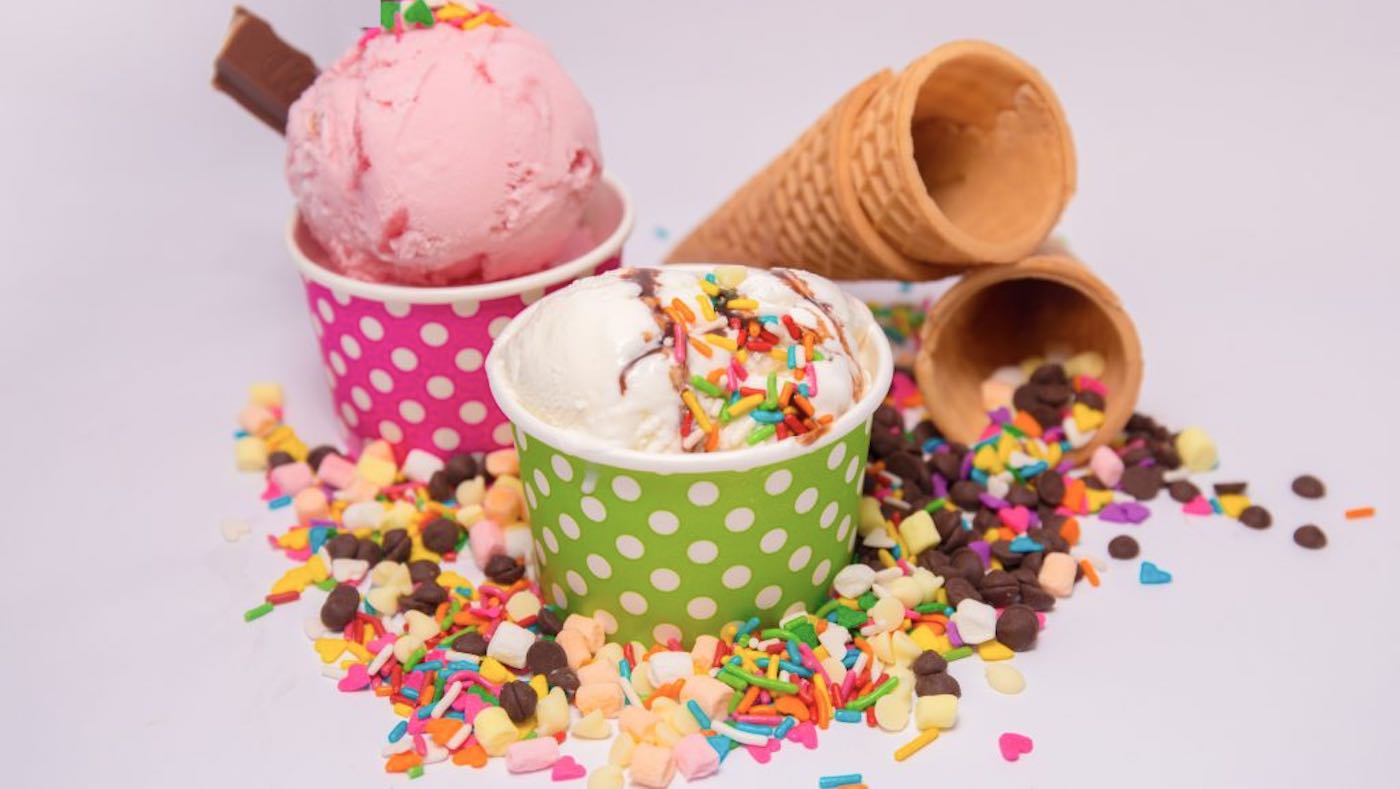 Two paper cups of ice cream with sprinkles surrounded by sprinkles and sugar cones