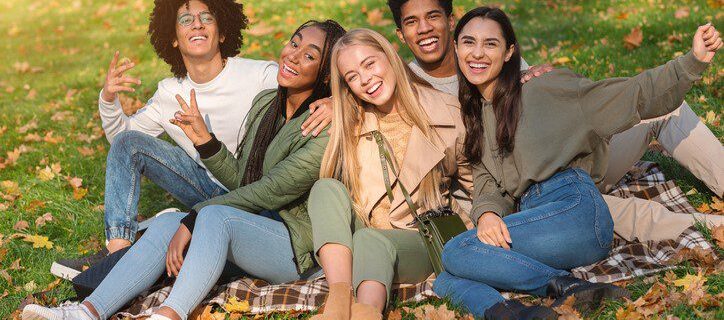 Cheerful Multiethnic Group of Teen Friends Having Fun In Autumn Park, sitting on the ground and posing at camera