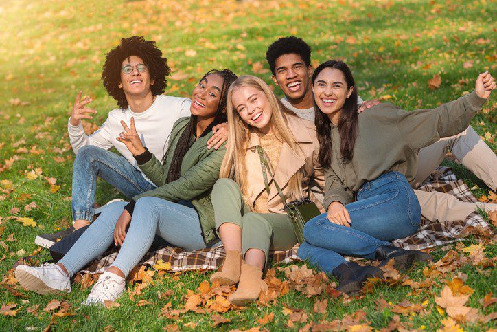 Cheerful Multiethnic Group of Teen Friends Having Fun In Autumn Park, sitting on the ground and posing at camera