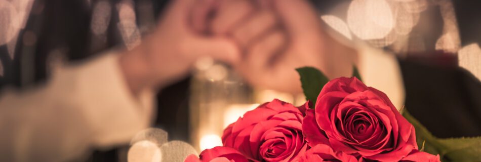 Couple at a candle light dinner date holding hands next to bouquet of red roses.