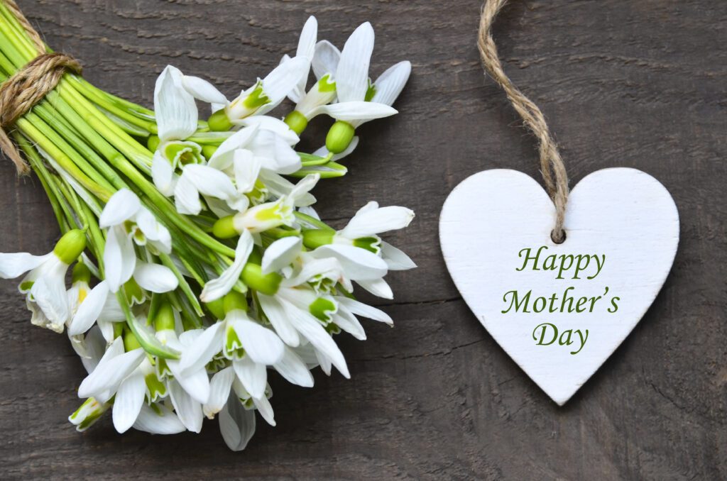 Happy Mother's Day greeting card with snowdrop spring flowers and decorative heart with text on old wooden table. Springtime holidays background. Selective focus.